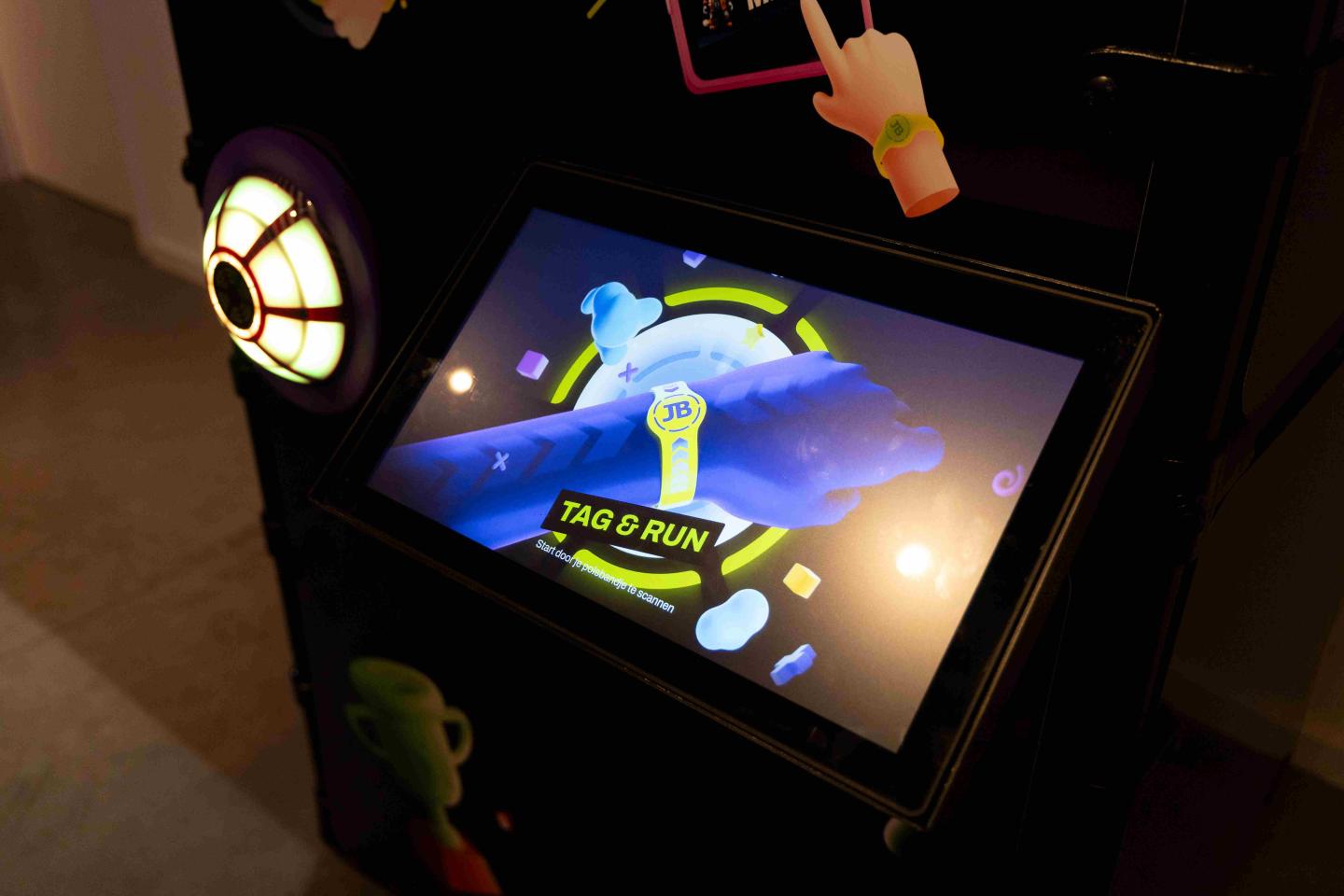 Close-up van het touchscreen van het JB TagSolutions spelsysteem met de animatie van een geel JB-polsbandje en de tekst “Tag & Run”, onderdeel van het interactieve registratiesysteem
