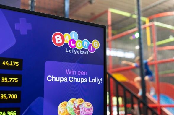 Close-up van een scherm in Ballorig Lelystad met een promotie om een lolly te winnen, met kleurrijk Ballorig-logo en speeltoestel op de achtergrond.