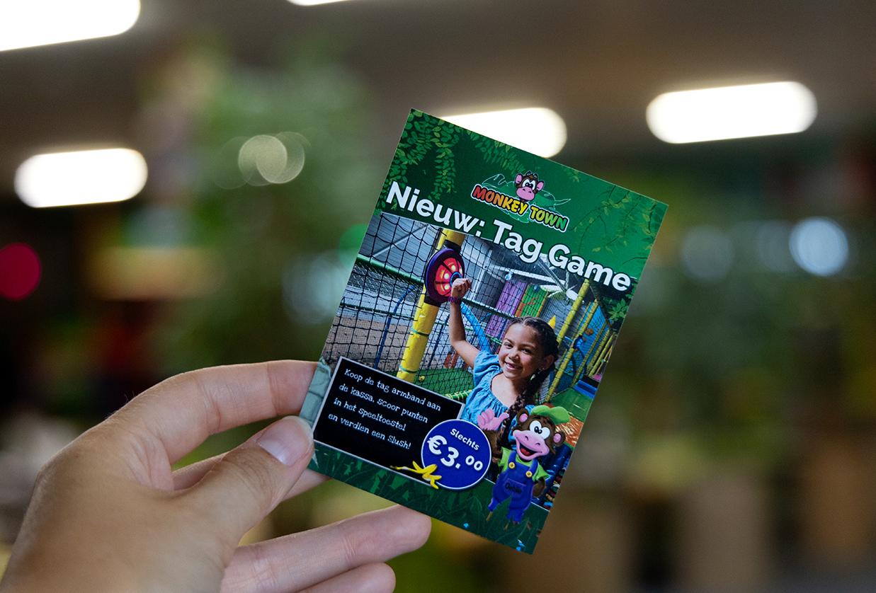 Hand houdt een flyer vast van Monkey Town met reclame voor de JB-TAG Game van JB Tag Solutions, waarin kinderen punten scoren met een tag-armband.